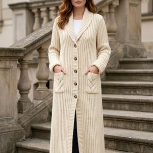Könnte beinhalten: Eine lange, cremefarbene Strickjacke im Rippenmuster mit Schalkragen, fünf braunen Knöpfen auf der Vorderseite und zwei aufgesetzten Taschen mit vertikalem Zopfmuster. Das Kleidungsstück reicht bis zum unteren Wadenbereich.