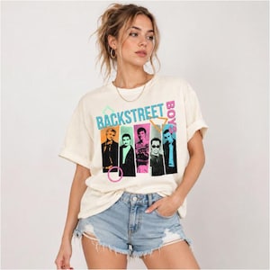 Pode incluir: Uma t-shirt de manga curta na cor creme com uma estampa gráfica dos Backstreet Boys. O design inclui cinco retratos individuais dos membros da banda em molduras retangulares coloridas, com o texto BACKSTREET BOYS em azul e rosa.