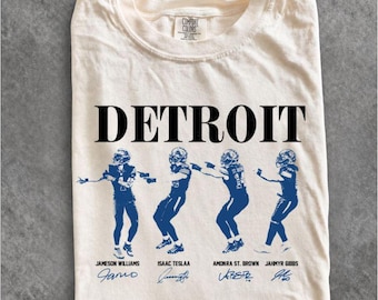 May include: A cream-colored t-shirt featuring the word DETROIT in black text above four blue illustrations of football players. Below each player are the names Jameson Williams, Isaac Teslaa, Amonra St. Brown, and Jahmyr Gibbs with signatures.