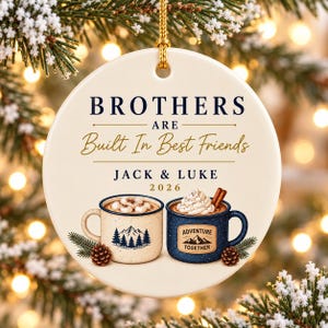 May include: A round white ceramic ornament with a gold hanging cord. The text reads BROTHERS ARE Built In Best Friends JACK & LUKE 2026. Below the text are two mugs, one cream with a mountain graphic and one blue with the text ADVENTURE TOGETHER.