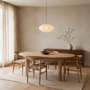 May include: An oval-shaped, off-white paper pendant light hangs above a light wood oval dining table. Four wooden chairs with woven seats surround the table, which holds a dark bowl. A wooden sideboard sits against the wall with a vase.
