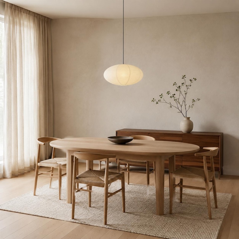 May include: An oval-shaped, off-white paper pendant light hangs above a light wood oval dining table. Four wooden chairs with woven seats surround the table, which holds a dark bowl. A wooden sideboard sits against the wall with a vase.