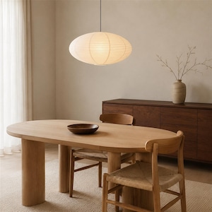 May include: An oval-shaped paper pendant light with horizontal ribbing hangs above an oval wooden dining table. The table features two thick cylindrical legs and is surrounded by wooden chairs with woven seats. A dark wooden bowl sits on the table.
