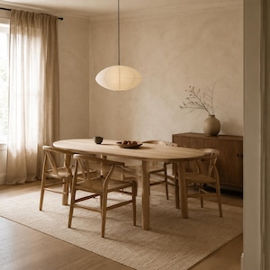 May include: An oval light wood dining table with four matching wooden chairs featuring woven seats. A white oval paper pendant light hangs above the table. A wooden sideboard with a ceramic vase and branches sits against the wall.