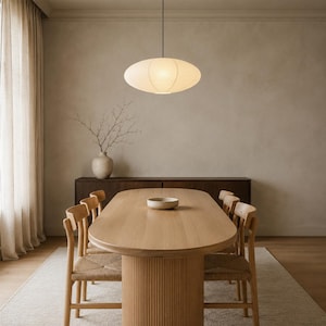 May include: An oval-shaped, off-white paper pendant light fixture hangs above a light wood oval dining table with a fluted base. Six wooden chairs with woven seats surround the table, which holds a small, shallow, light-colored bowl.