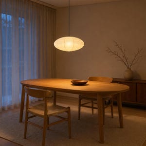 Puede incluir: Una lámpara colgante de papel de forma ovalada cuelga sobre una mesa de comedor de madera. La mesa es ovalada con cuatro patas y está acompañada por dos sillas de madera con asientos trenzados. Un cuenco de madera oscura se sitúa en el centro.