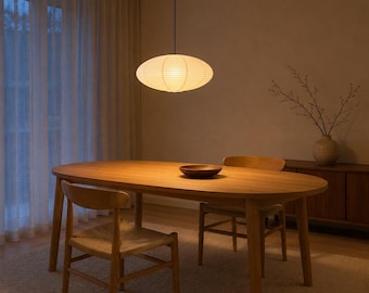 Peut inclure: Une suspension en papier de forme ovale est suspendue au-dessus d'une table à manger en bois. La table est ovale avec quatre pieds et est accompagnée de deux chaises en bois avec des assises tressées. Un bol en bois sombre est posé au centre.