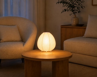 May include: A round, off-white paper lantern table lamp with a ribbed texture and a warm, glowing light. The lamp features a simple, thin metal tripod base that elevates the spherical shade slightly above the surface of the wooden table.
