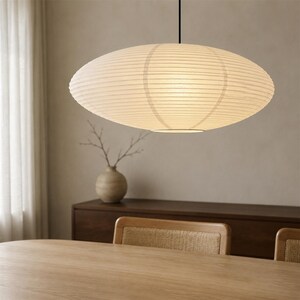 May include: A large, oval-shaped pendant light with a ribbed, off-white paper shade. The fixture is suspended by a thin black cord from the ceiling, emitting a soft, diffused glow through the textured material of the rounded lantern structure.