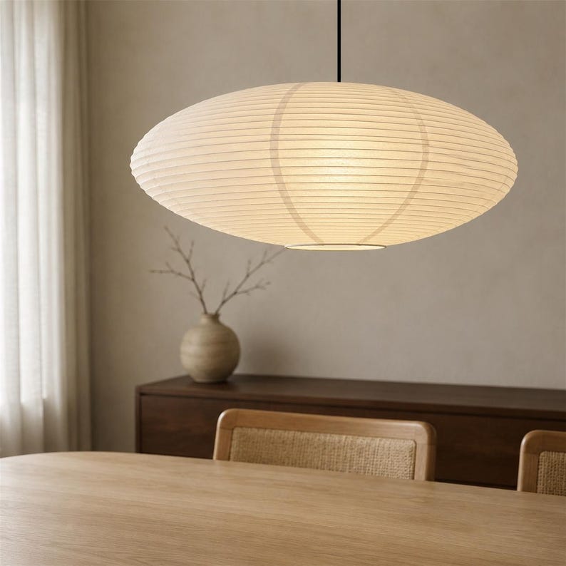 May include: A large, oval-shaped pendant light with a ribbed, off-white paper shade. The fixture is suspended by a thin black cord from the ceiling, emitting a soft, diffused glow through the textured material of the rounded lantern structure.