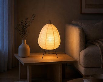 May include: A table lamp with a cream-colored, ribbed paper shade shaped like a teardrop. The lamp features a thin black metal frame with three slender legs that support the shade. It emits a warm, soft glow, illuminating the surrounding area.
