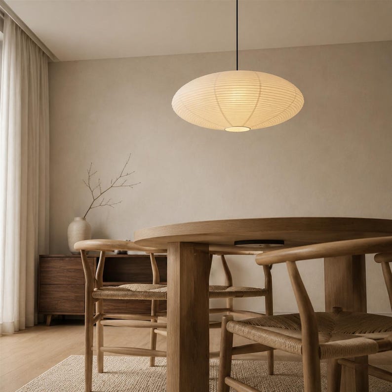May include: A saucer-shaped pendant light with a ribbed off-white paper shade suspended by a thin black cord. The light fixture emits a soft, warm glow, featuring a circular design that hangs centrally above a wooden dining table and chairs.