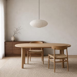 May include: An oval light wood dining table with four matching chairs featuring woven seats. A large, elliptical, off-white paper pendant light hangs above the table. A dark wood sideboard sits against the wall with a vase containing bare branches.