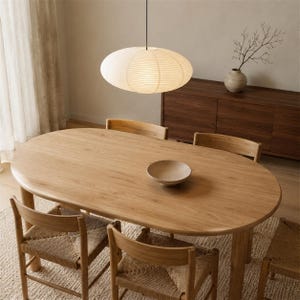 May include: An oval light wood dining table with four matching chairs featuring woven seats. A large, round, off-white paper pendant light hangs above the table. A small, shallow, light-colored bowl sits in the center of the wooden tabletop.