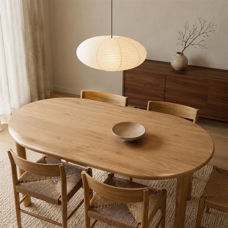 May include: An oval light wood dining table with four matching chairs featuring woven seats. A large, round, off-white paper pendant light hangs above the table. A small, shallow, light-colored bowl sits in the center of the wooden tabletop.