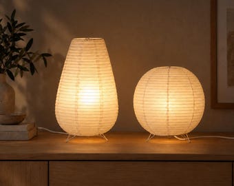 May include: Two off-white paper table lamps with a ribbed texture and warm internal illumination. The left lamp has a tall, tapered, pear-like shape, while the right lamp is spherical. Both lamps feature thin, metallic tripod legs and white cords.