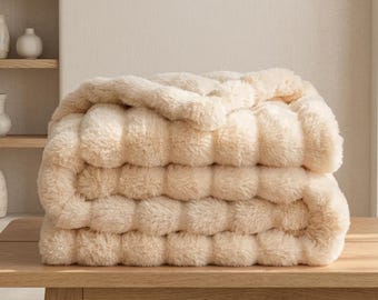 May include: A plush, cream-coloured faux fur throw blanket folded into a thick, layered stack. The fabric features a dense, shaggy texture with a soft, uniform appearance, showcasing a warm, neutral tone throughout the entire folded textile piece.