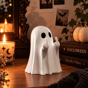 Op de afbeelding: Een wit spookfiguurtje met twee zwarte ovale ogen, afgebeeld met beide armen omhoog en de middelvingers uitgestoken. Het gladde, mat afgewerkte spook heeft een gedrapeerd, lakenachtig uiterlijk met verticale plooien aan de onderkant.