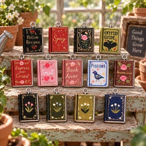 May include: A collection of twelve rectangular book-shaped metal charms with silver-toned loops. Each charm features unique cover art, including titles like Potion Recipes, Spicy, Crytid Guide, Express your Crues, Smut, Book lover, and Pigeons, alongside floral designs.