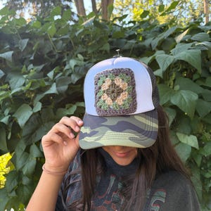 May include: A trucker cap featuring a white front panel adorned with a square, multi-coloured crochet patch in shades of brown, tan, and sage green. The brim displays a camouflage pattern, and the back consists of a black mesh material.