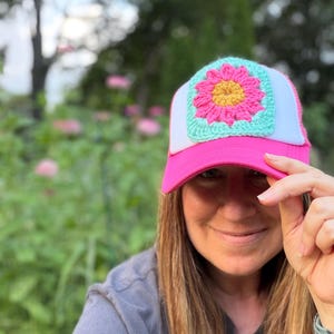 Könnte beinhalten: Eine Trucker-Cap mit einem leuchtend pinken Schirm und einem weißen Netzrücken. Das Vorderteil ist mit einem quadratischen Häkelaufnäher verziert, der eine leuchtend pinke Blume mit gelber Mitte auf hellminzgrünem Grund zeigt.
