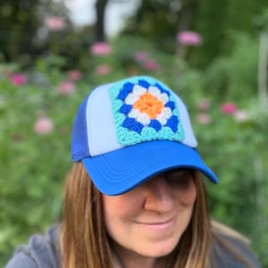 May include: A blue trucker hat featuring a white front panel adorned with a square, multi-coloured crochet granny square patch. The patch displays a bright orange centre surrounded by layers of white, dark blue, and light turquoise yarn stitches.