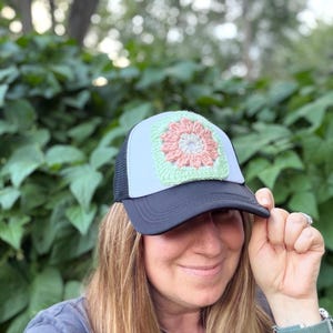 Könnte beinhalten: Eine Trucker-Cap mit schwarzem Schirm und schwarzem Netzrücken sowie einer weißen Vorderseite. Die Vorderseite ist mit einem quadratischen, hellgrünen Häkelaufnäher verziert, der ein zentrales Blumenmotiv mit zartrosa Blütenblättern zeigt.