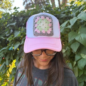 Könnte beinhalten: Eine hellrosa und weiße Trucker-Cap mit einem quadratischen Häkelaufnäher auf der Vorderseite. Das Motiv besteht aus konzentrischen Garnschichten in den Farben Creme, Hellgrün, Hellrosa und Dunkelgrau.