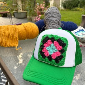 Könnte beinhalten: Eine grüne und weiße Trucker-Cap mit einem quadratischen, mehrfarbigen Häkelaufnäher auf der Vorderseite. Der Aufnäher besteht aus konzentrischen Schichten aus hellblauem, pinkem, schwarzem und grünem Garn in einem klassischen Granny-Square-Muster.