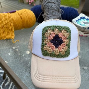 Könnte beinhalten: Eine beigefarbene Trucker-Cap mit weißem Vorderteil, das mit einem quadratischen Häkelaufnäher verziert ist. Der Aufnäher zeigt ein mehrfarbiges Blumenmuster in Dunkelblau, Altrosa, Beige und Olivgrün, das mittig auf dem weißen Stoff sitzt.