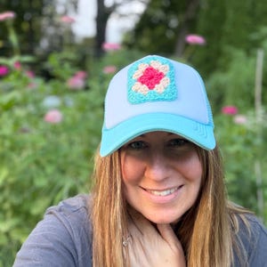 Könnte beinhalten: Eine hellblaue und weiße Trucker-Cap mit einem quadratischen Häkelaufnäher auf der Vorderseite. Der Aufnäher besteht aus einer leuchtend pinken Mitte, umgeben von einem cremefarbenen Rand und einer hellblauen Umrandung, passend zum Schirm.