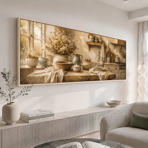 May include: A rectangular wall art print depicting a sunlit kitchen scene with a wooden table covered by a draped cloth. The table holds ceramic bowls, a pitcher, and a large vase filled with dried flowers near a window. The background shows shelves.