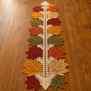 May include: A long, narrow table runner featuring a central cream-coloured crocheted spine with alternating leaf-shaped motifs in shades of deep red, burnt orange, golden yellow, and olive green, creating a symmetrical, textured autumn-inspired design.