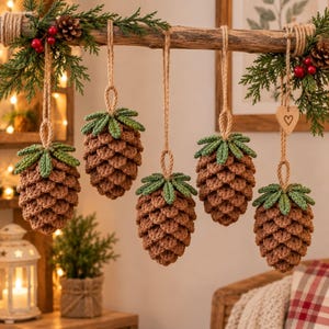 May include: Five crocheted pinecone ornaments hanging from a horizontal wooden branch. Each ornament features a brown textured body with green crocheted leaves at the top, suspended by a tan twine loop. A small wooden heart charm hangs on the right.