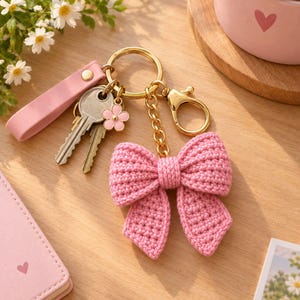 May include: A gold-toned metal keyring featuring a pink crocheted bow charm, a small pink flower charm, and a pink leather-like strap. The keyring includes a split ring and a lobster clasp, with two silver-coloured metal keys attached to the ring.