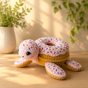 May include: A crocheted turtle plush toy featuring a shell shaped like a donut with pink frosting and colorful sprinkles. The head and flippers are also pink with sprinkles, while the underside of the shell and flippers are a solid golden brown.