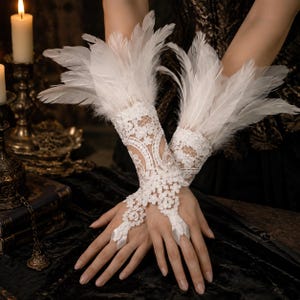 May include: A pair of white lace fingerless gloves featuring intricate floral embroidery and long, soft white feathers attached at the wrist. The gloves are designed to cover the back of the hand and forearm, securing around the middle finger with a loop.