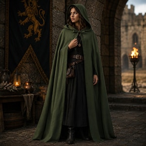 May include: A floor-length olive green hooded cloak featuring a simple tie closure at the neck. The garment is worn over a dark long-sleeved dress with a brown leather belt and a small attached pouch, creating a full-length layered appearance.