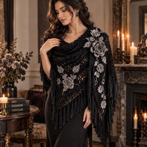 May include: A black triangular crochet shawl featuring intricate openwork patterns, long fringe along the edges, and three-dimensional grey floral appliqués with leaves attached to the fabric. The shawl is draped over a plain black garment.