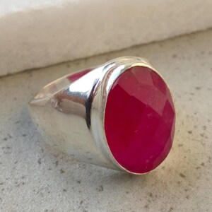 Op de afbeelding: Een zilveren ring met een grote, ovaalvormige, gefacetteerde dieproze edelsteen gezet in een gepolijste metalen band. De steen heeft een getextureerd, veelzijdig oppervlak dat licht reflecteert en de band is glad en metaalachtig met een glanzende afwerking.