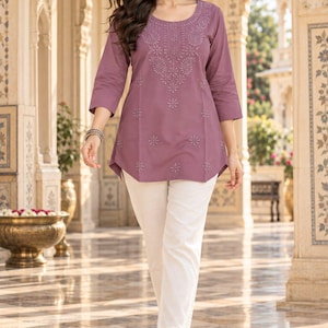 May include: A mauve tunic featuring intricate tonal embroidery around the scoop neckline and scattered floral motifs across the front, paired with straight-leg off-white trousers. The tunic has three-quarter length sleeves and a curved hemline.