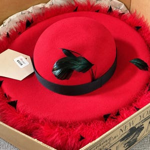 May include: A bright red felt hat with a wide brim trimmed in red feathers. The hat features a black ribbon band around the crown, accented with dark green feathers. A hexagonal tag with the number 6119 is attached to the brim of the hat.