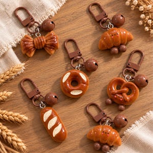 May include: A collection of six brown food-themed keychains featuring a bow, two croissants, a donut, a pretzel, and a baguette. Each keychain includes a brown metal lobster clasp and a small round bell, with some items displaying simple faces.