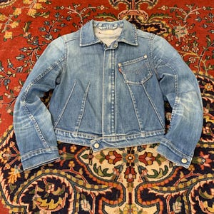 Può includere: Una giacca in denim azzurro con colletto a punta, maniche lunghe e una tasca singola sul petto con una piccola etichetta rossa. La giacca presenta una chiusura frontale con bottoni, cuciture verticali visibili e un orlo corto con un bottone alla base.