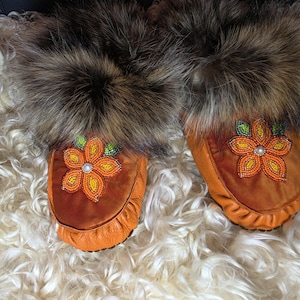 May include: A pair of tan leather moccasins featuring a thick, dark brown fur trim around the opening. Each moccasin is decorated with a beaded floral design on the top, showcasing orange and yellow petals with a small white bead in the centre.