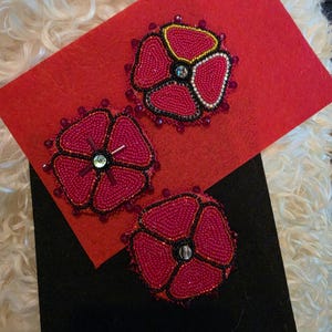 Peut inclure: Trois écussons perlés circulaires en forme de fleur, composés de perles de rocaille rose vif avec des contours noirs. Chaque écusson présente un accent métallique central et une bordure de petites perles sombres sur fond de feutrine rouge et noire.