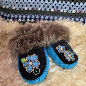 May include: A pair of black and bright blue moccasins featuring a brown fur trim around the opening. The top of each moccasin is decorated with intricate blue beaded flower patterns with small green leaves and a central white bead detail.