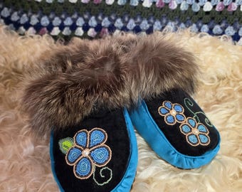 May include: A pair of black and bright blue moccasins featuring a brown fur trim around the opening. The top of each moccasin is decorated with a beaded floral design in shades of blue, green, and white, showcasing intricate beadwork.