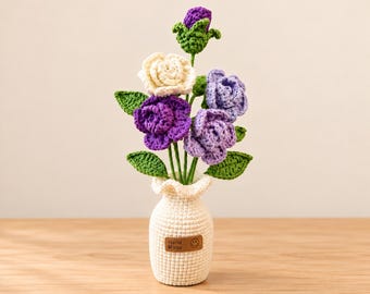 May include: A crochet flower arrangement featuring a cream rose, two purple roses, and a small purple bud with green leaves. The flowers are held in a cream-colored crochet vase with a ruffled top and a brown tag labeled Hand Made.