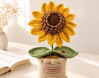 May include: A crocheted sunflower with bright yellow petals and a textured brown center, featuring two green leaves and a small red ladybug accent. The flower is potted in a beige crocheted base with a leather-like tag labeled HANDMADE Limited edition.